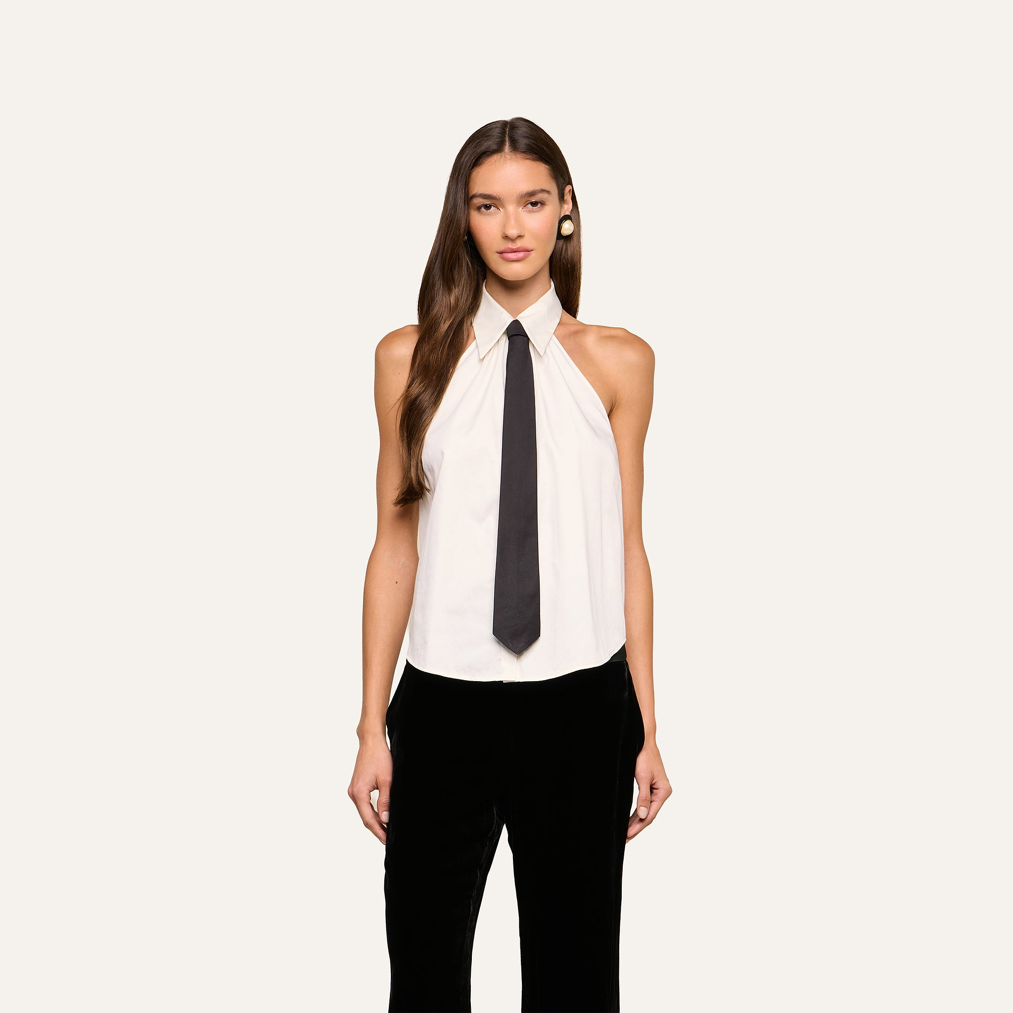 A woman stands facing forward against a plain light background, wearing the Wren Neck-Tie Top—a sleeveless white halter button-down top with a black necktie—paired with black pants.