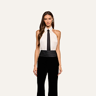 A woman with long brown hair wears the Wren Neck-Tie Top— a sleeveless, halter black and white button-down piece featuring a black tie detail—paired with wide-leg pants, standing against a plain light background.