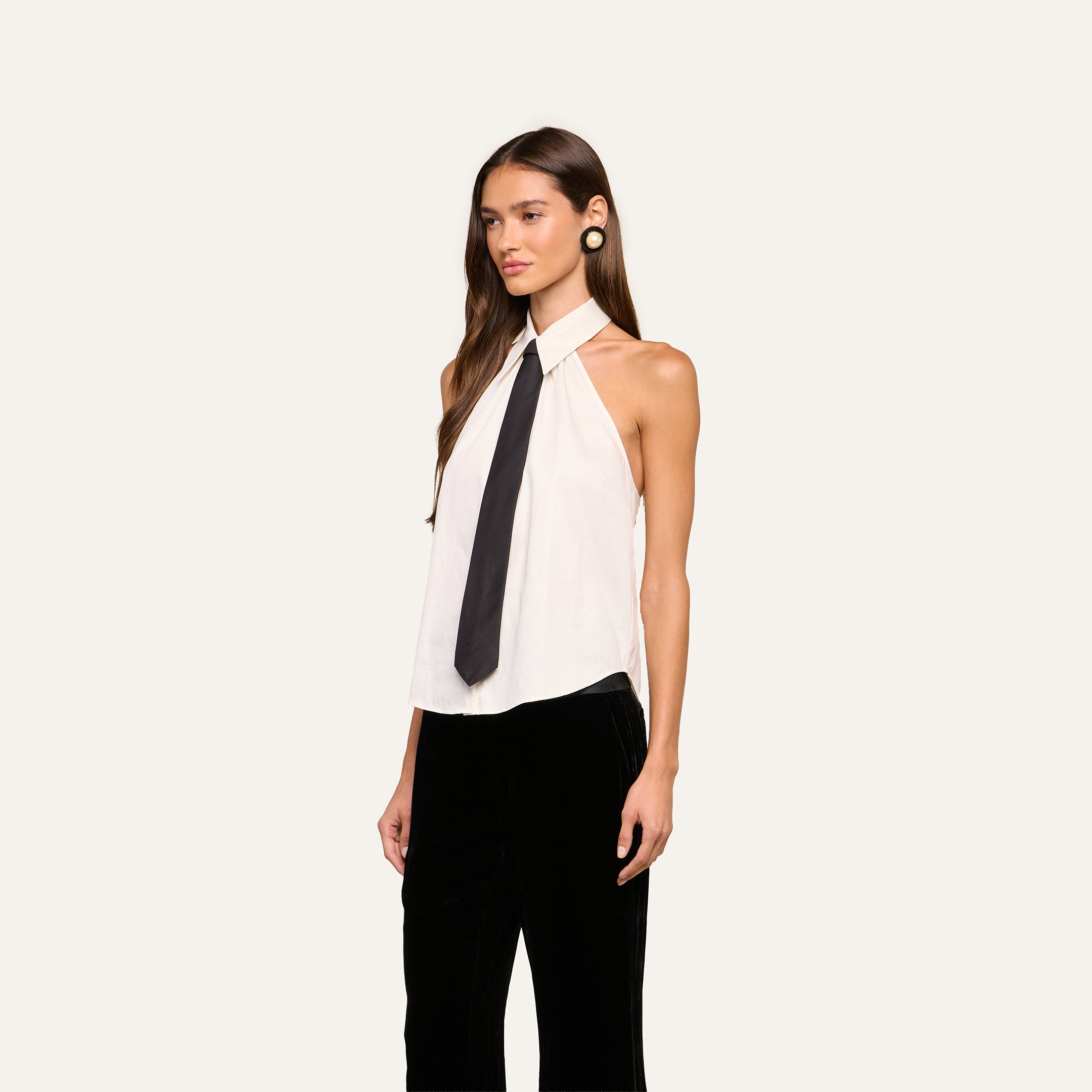 A woman stands wearing the Wren Neck-Tie Top—a sleeveless white halter button-down with a collar and black necktie—paired with black pants. She has long brown hair, large round earrings, and is posed against a plain, light-colored background.