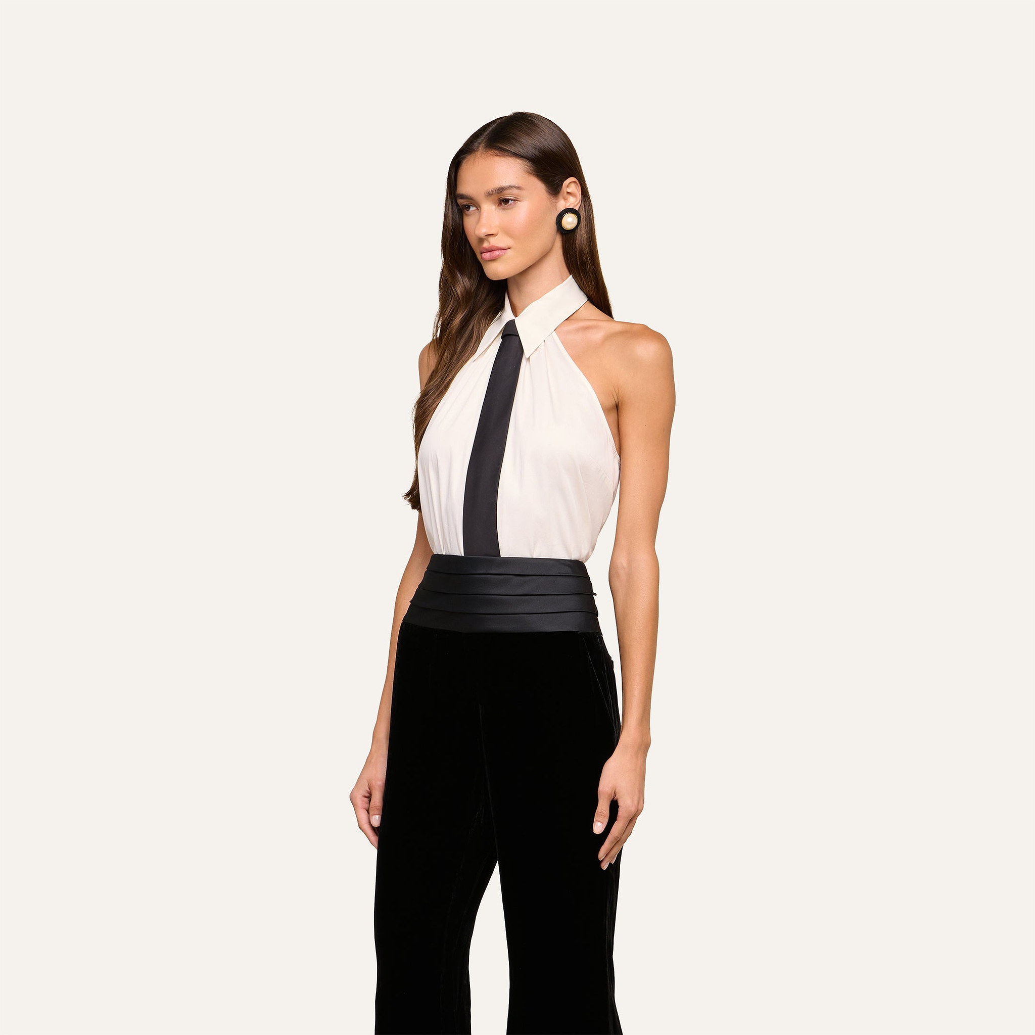 A woman wearing a sleeveless white Wren Neck-Tie Top with a black tie, paired with black high-waisted pants and gold earrings, stands against a plain light background.