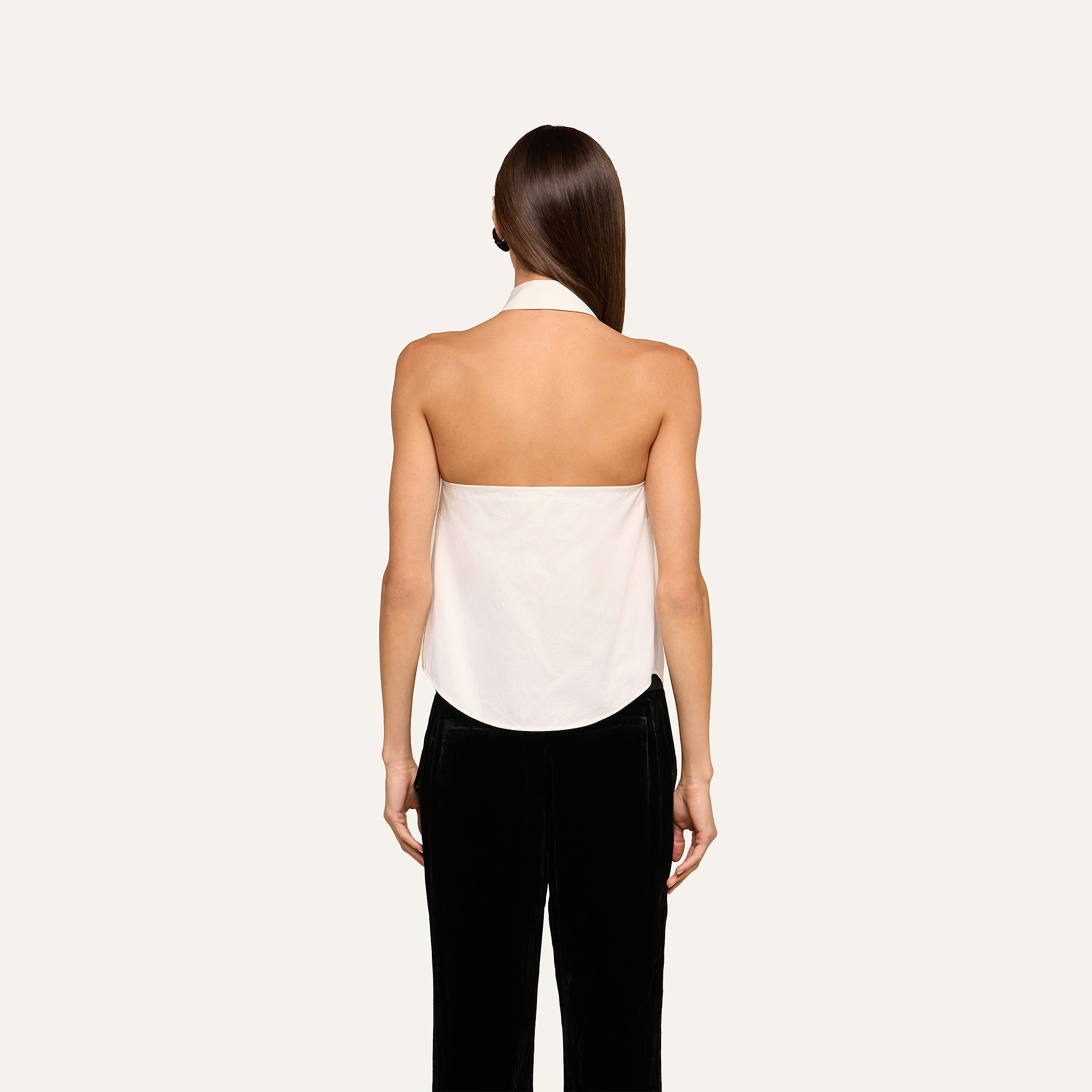 A woman stands facing away against a plain light background, wearing the Wren Neck-Tie Top—a white halter button-down style—paired with black pants. Her straight brown hair is down and her arms are at her sides.