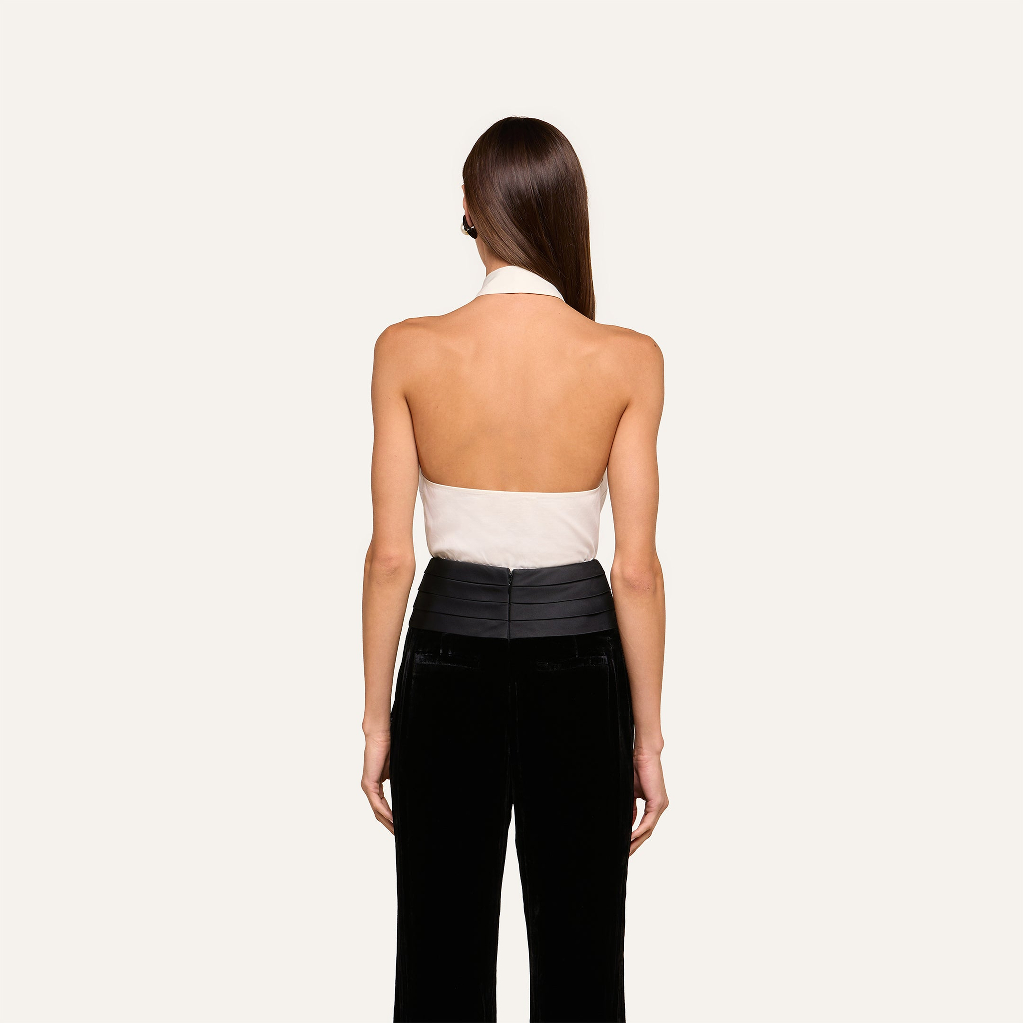 A woman with straight brown hair stands facing away, wearing a white Wren Neck-Tie Top and black high-waisted pants against a plain light background.