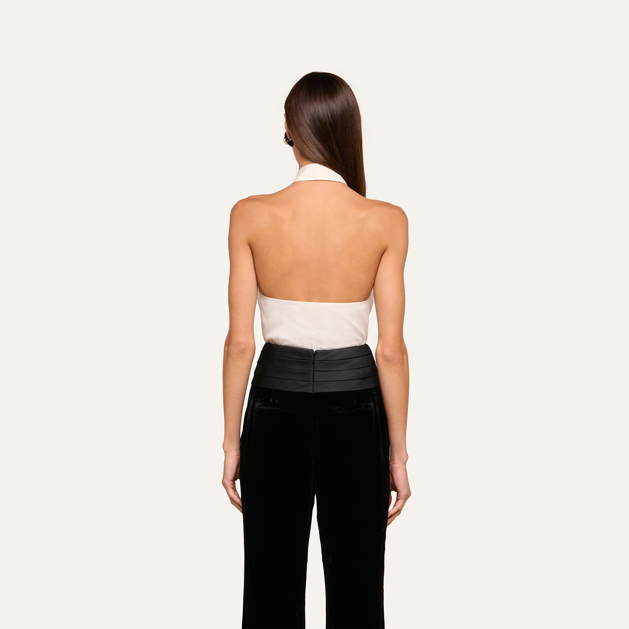A woman with straight brown hair stands facing away, wearing a white Wren Neck-Tie Top and black high-waisted pants against a plain light background.