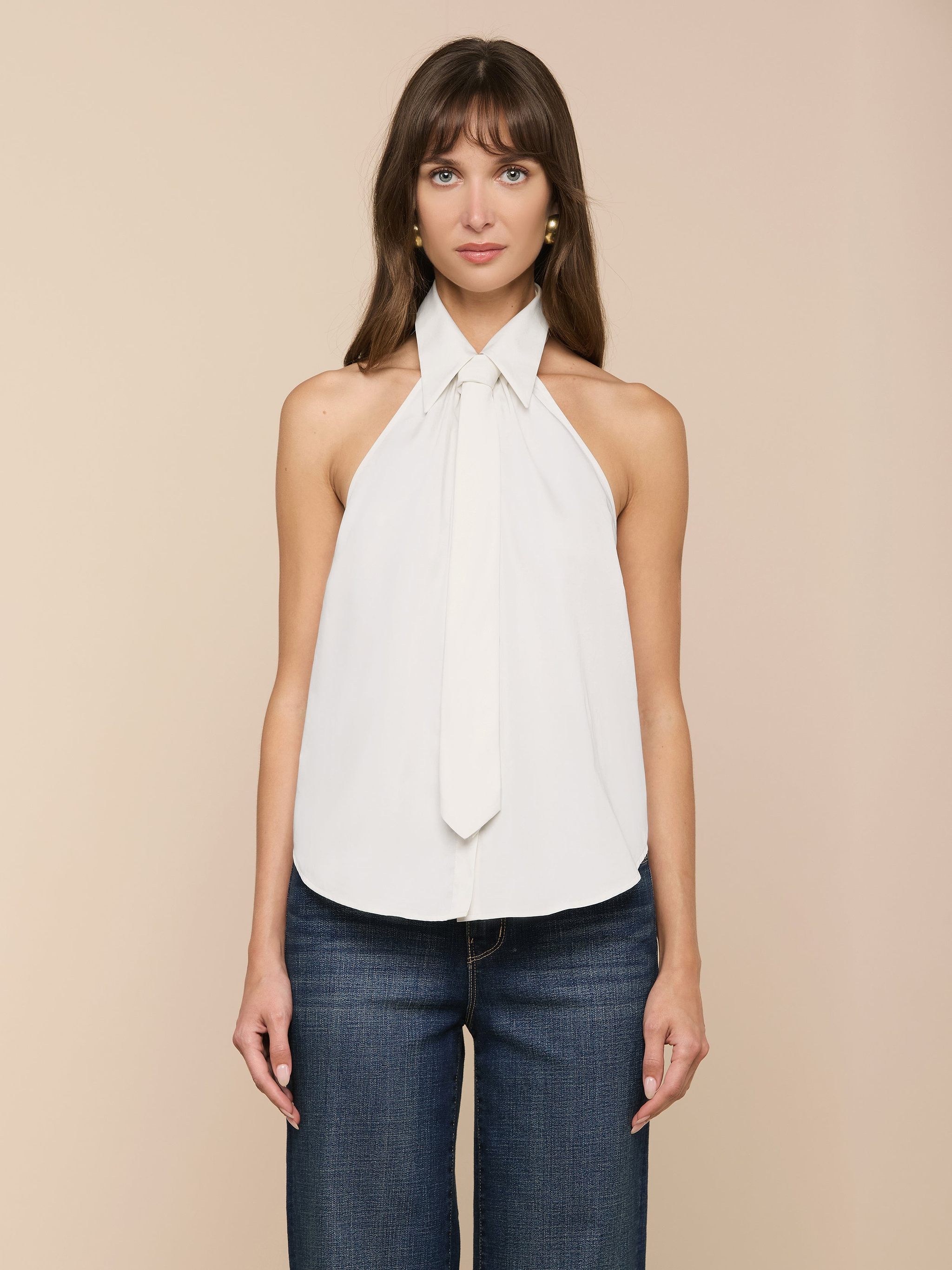 Eine Frau mit langen braunen Haaren trägt das Wren Poplin Top, ein ärmelloses weißes Halstuch mit einer Krawattenverzierung aus weicher Satin-Popeline, gepaart mit dunkelblauen Jeans, vor einem schlichten beigen Hintergrund.