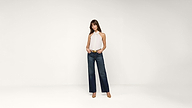 A woman stands in front of a plain white background, wearing a white halter top, dark wide-leg jeans, and tan heels. She looks forward with one hand on her hip.