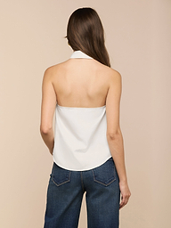 Eine Frau mit langen braunen Haaren wird von hinten gezeigt. Sie trägt ein weißes Zaunkönig-Popeline-Top in einer Neckholder-Silhouette und blaue Jeans vor einem schlichten beigen Hintergrund.
