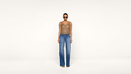 A woman wearing sunglasses, a sleeveless patterned top, and wide-leg blue jeans stands against a plain light background, facing forward.