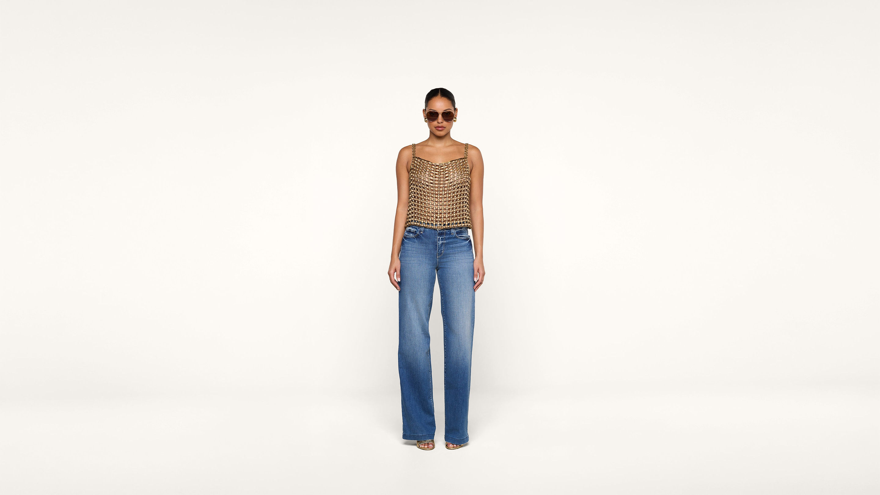 A woman wearing sunglasses, a sleeveless patterned top, and wide-leg blue jeans stands against a plain light background, facing forward.