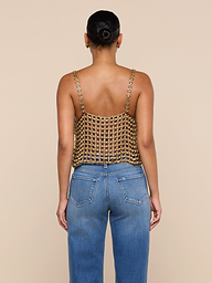 A woman stands facing away, wearing the Joanette Embellished Tank—a gold metallic chainmail-style, hand-woven top with chain straps—paired with blue jeans against a plain off-white background.