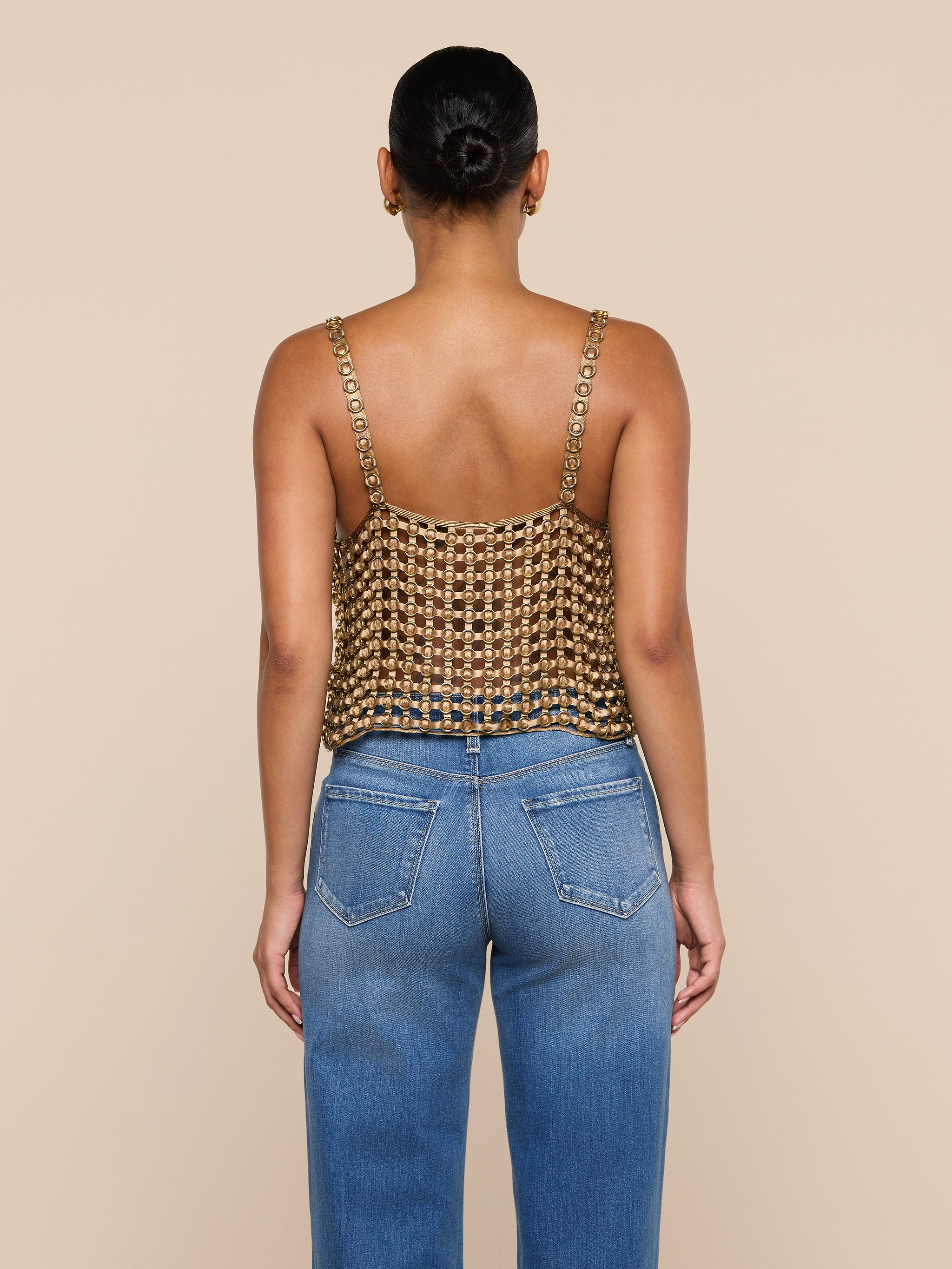 A woman stands facing away, wearing the Joanette Embellished Tank—a gold metallic chainmail-style, hand-woven top with chain straps—paired with blue jeans against a plain off-white background.