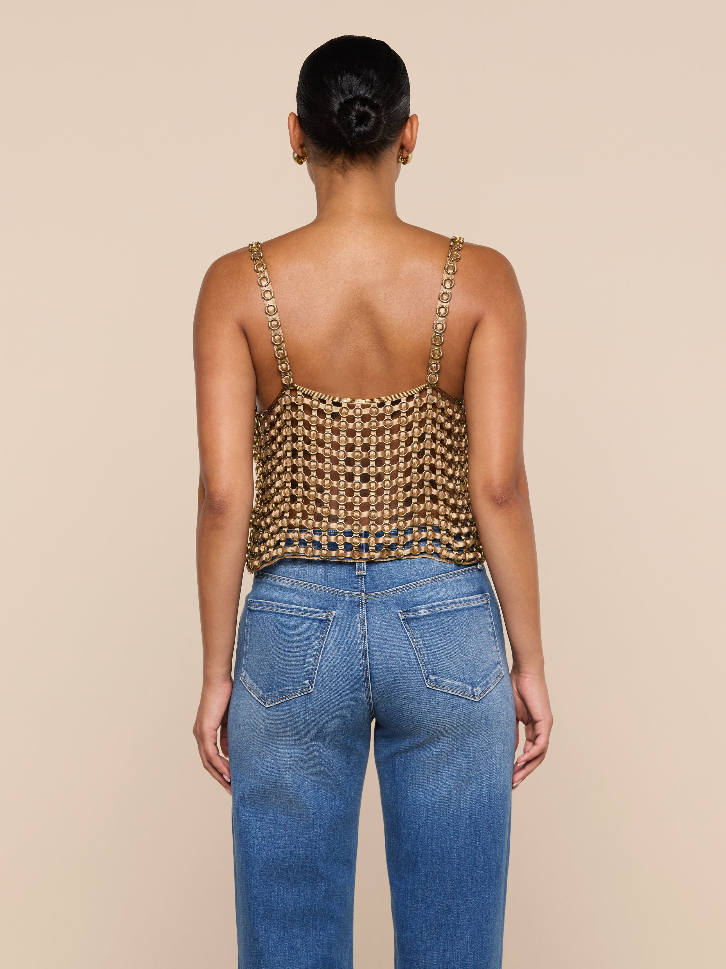 A woman stands facing away, wearing the Joanette Embellished Tank—a gold metallic chainmail-style, hand-woven top with chain straps—paired with blue jeans against a plain off-white background.
