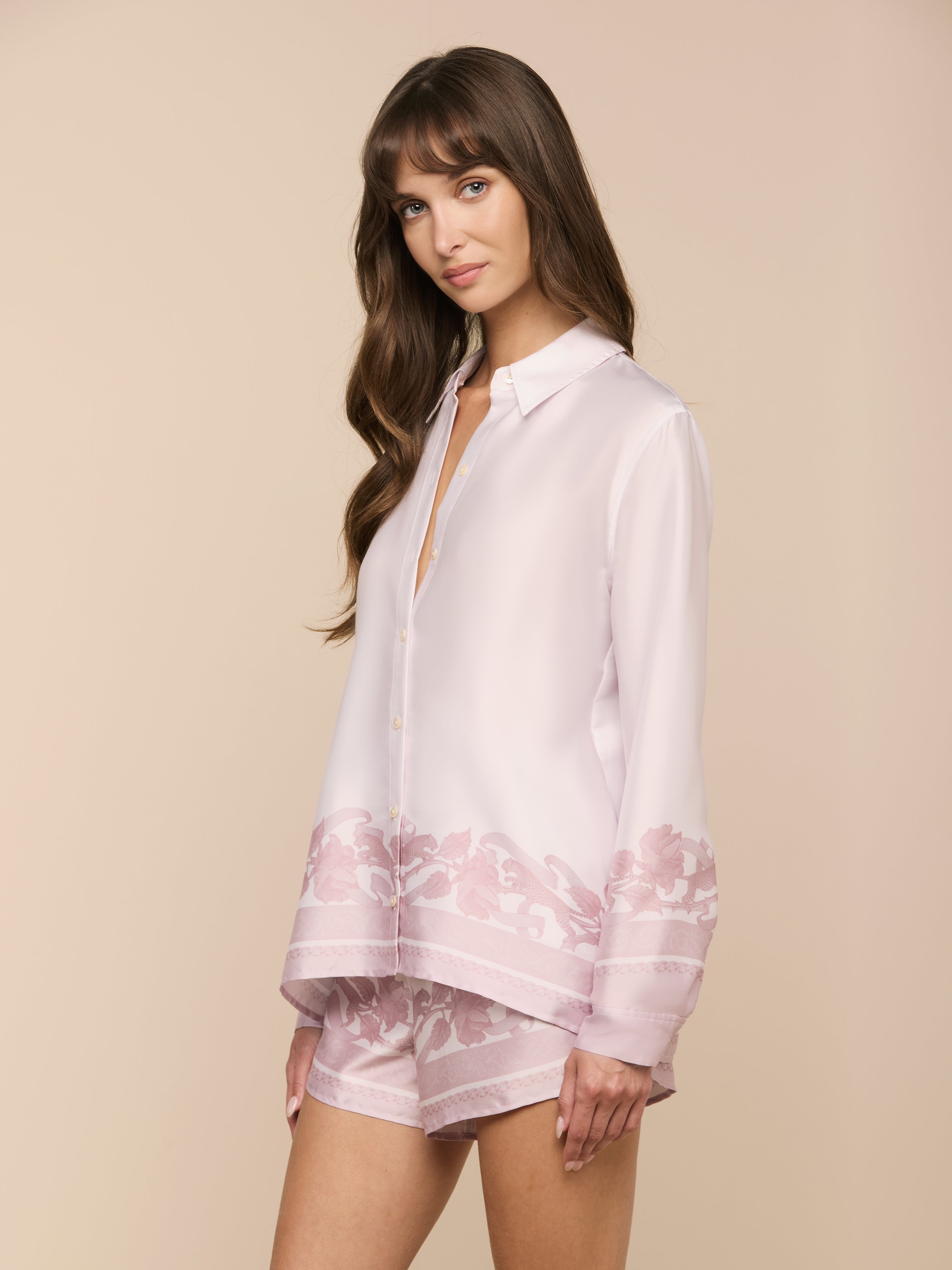 A woman with long brown hair wears the Judy Blouse, a light pink button-down top featuring floral patterns on the hem, paired with matching pink shorts, standing against an off-white background.