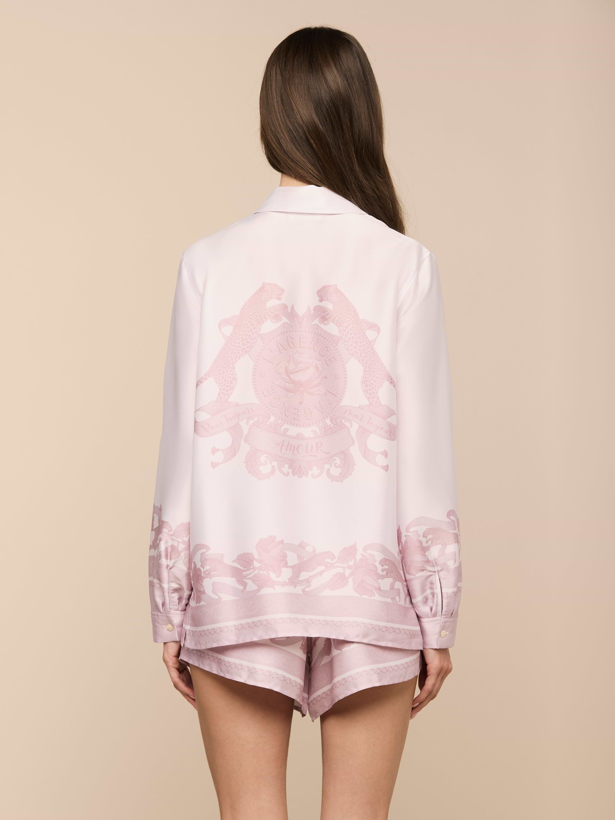Wearing the Judy Blouse, a woman with long brown hair stands with her back to the camera against a plain, light background. The light pink button-down pajama set features decorative patterns and a crest on the back.