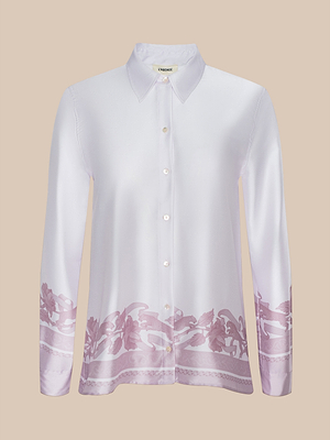 The Judy Blouse is a long-sleeve white button-down with a pointed collar, light pink floral accents on the bottom and sleeve cuffs, made from soft poly twill and shown on a beige background.