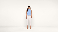 A woman with long brown hair stands against a plain white background, wearing a light blue sleeveless top, white pants, and beige heels. She faces forward with a neutral expression and her arms relaxed by her sides.