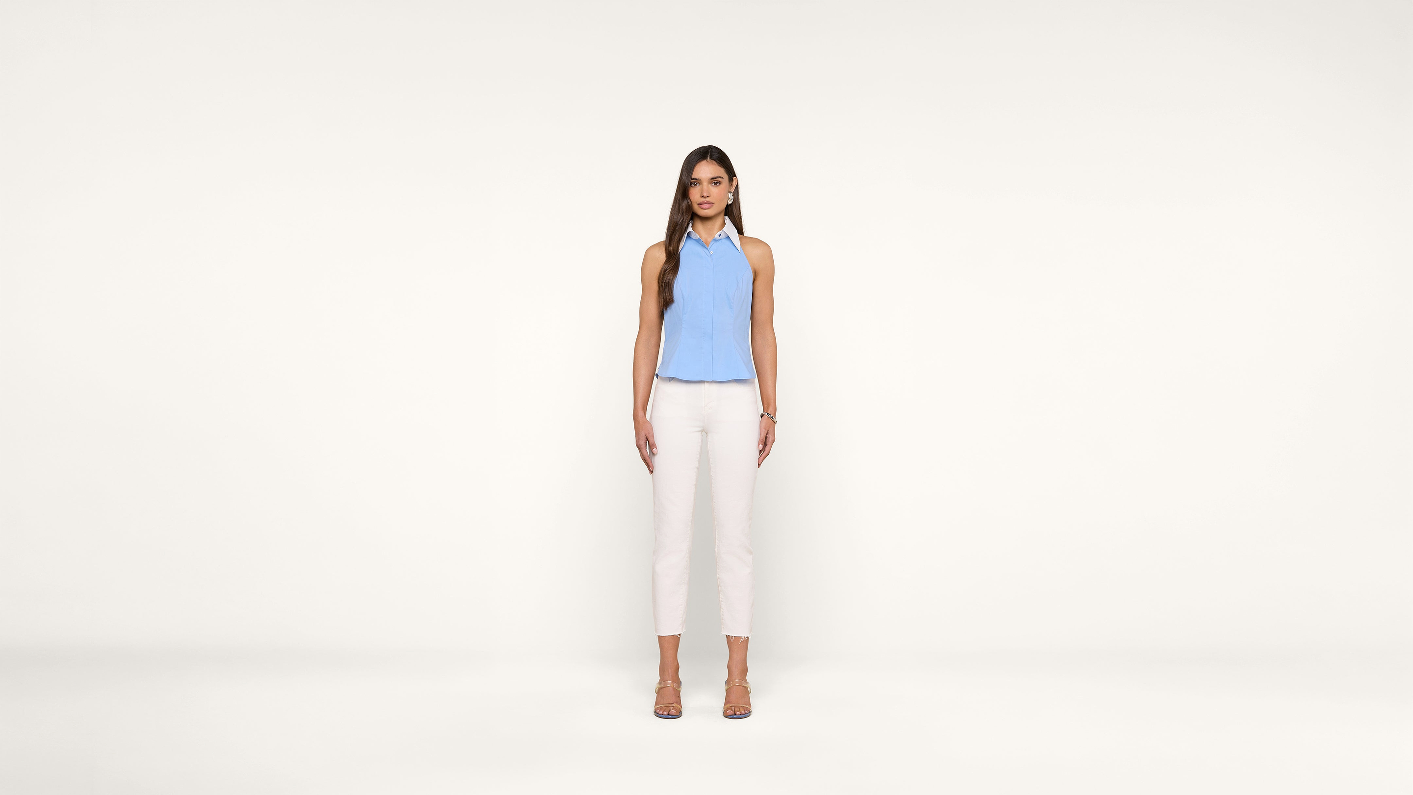 A woman with long brown hair stands against a plain white background, wearing a light blue sleeveless top, white pants, and beige heels. She faces forward with a neutral expression and her arms relaxed by her sides.
