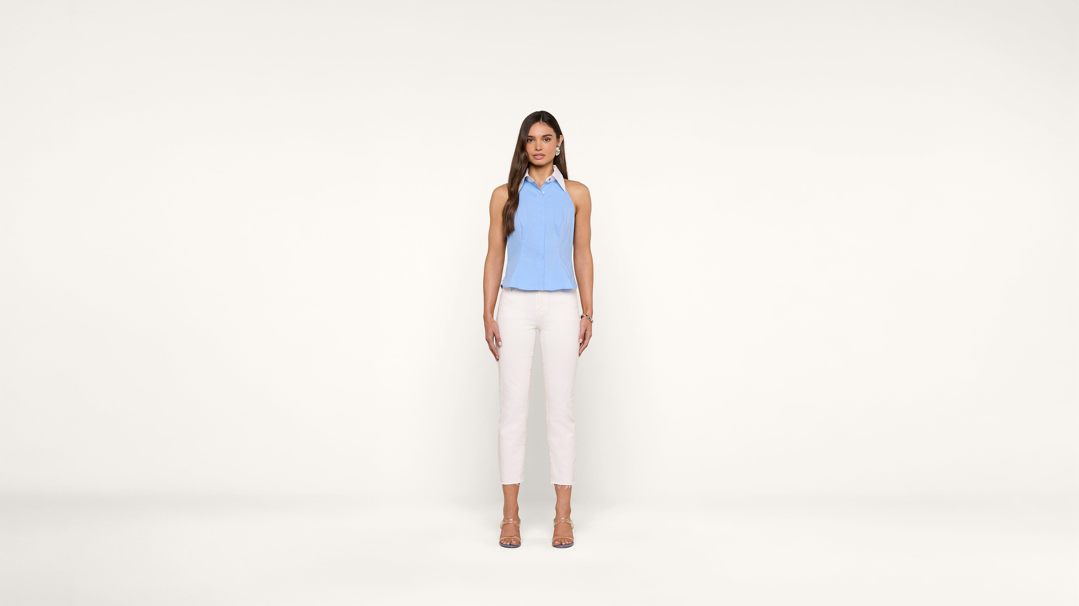 A woman with long brown hair stands against a plain white background, wearing a light blue sleeveless top, white pants, and beige heels. She faces forward with a neutral expression and her arms relaxed by her sides.