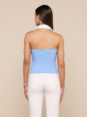 A woman with long brown hair wears the Rina Poplin Halter Top, a light blue strapless organic cotton-blend top with a white halter neck, paired with high-waisted white pants and silver hoop earrings and bracelet.
