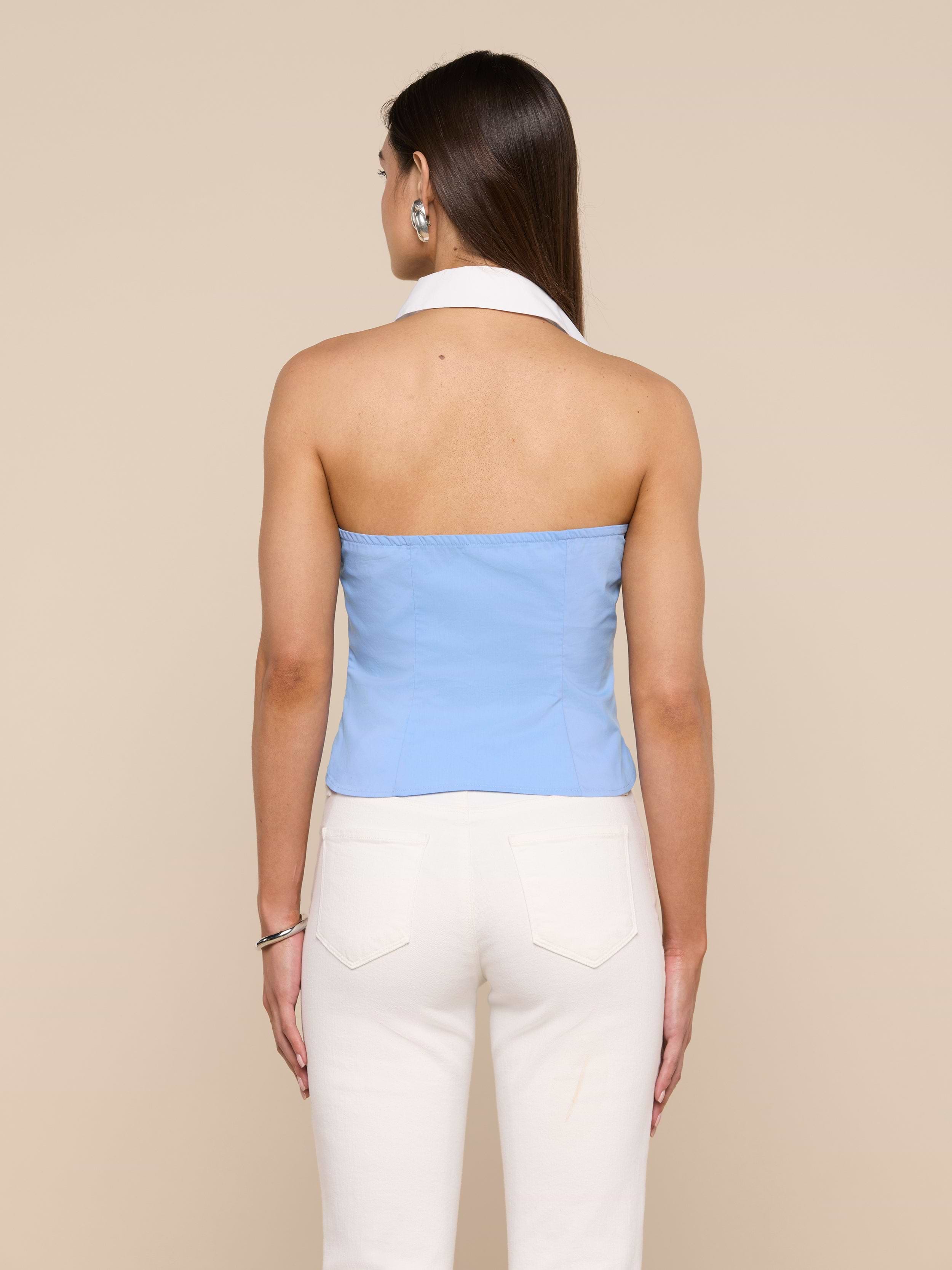 A woman stands with her back to the camera, wearing the light blue Rina Poplin Halter Top and white pants against an off-white background.