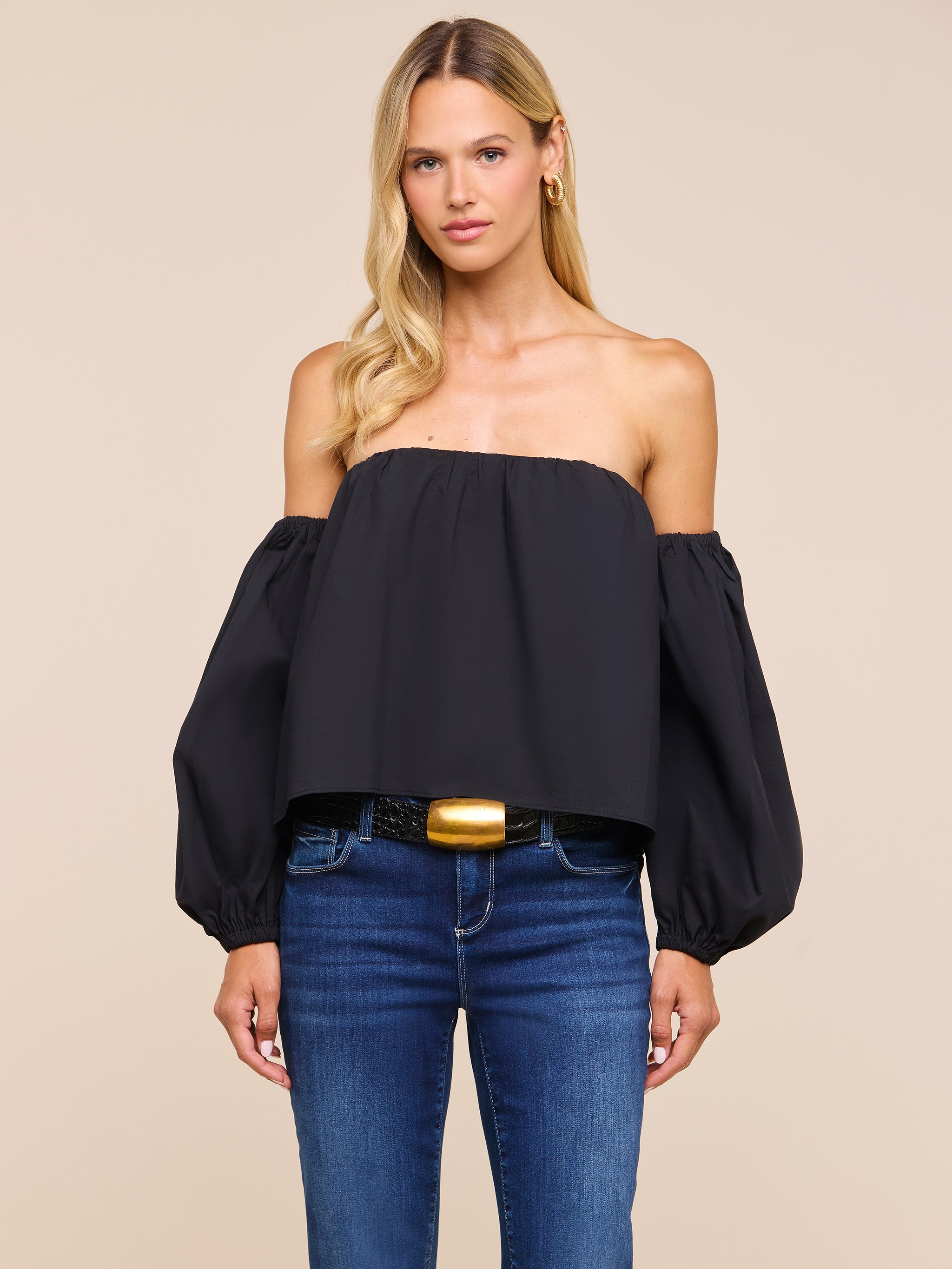A woman with long blonde hair wears the Sarai Off-The-Shoulder Top, styled in black and paired with blue jeans, standing against a plain light background.