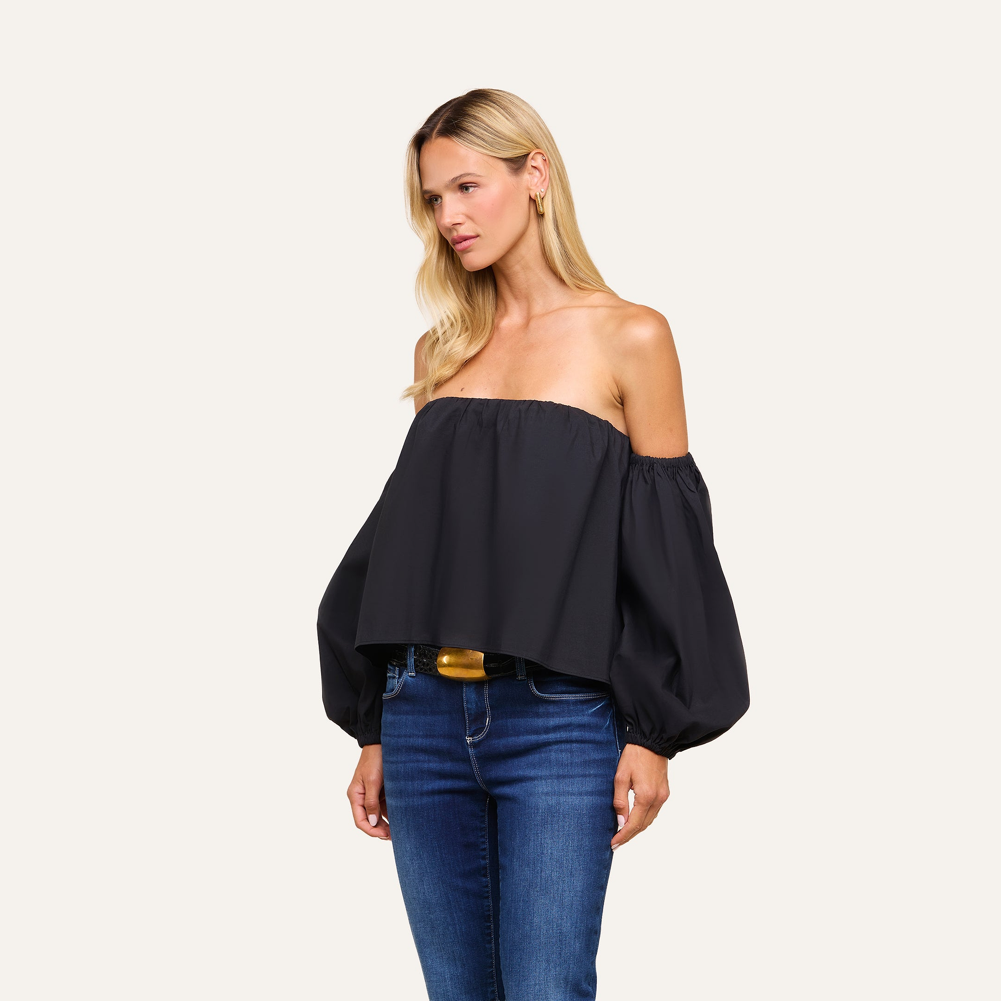 Wearing the Sarai Off-The-Shoulder Top—a black, long balloon-sleeved piece crafted from an organic cotton-blend—a woman pairs it with blue jeans and poses against a plain light background, gazing to the side. The top’s fabric ensures both comfort and style.