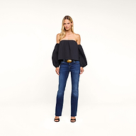 A woman stands against a white background wearing the Sarai Off-The-Shoulder Top—an organic cotton-blend black piece featuring puffed sleeves—paired with blue jeans, black open-toe heels, and a wide belt with a gold buckle.