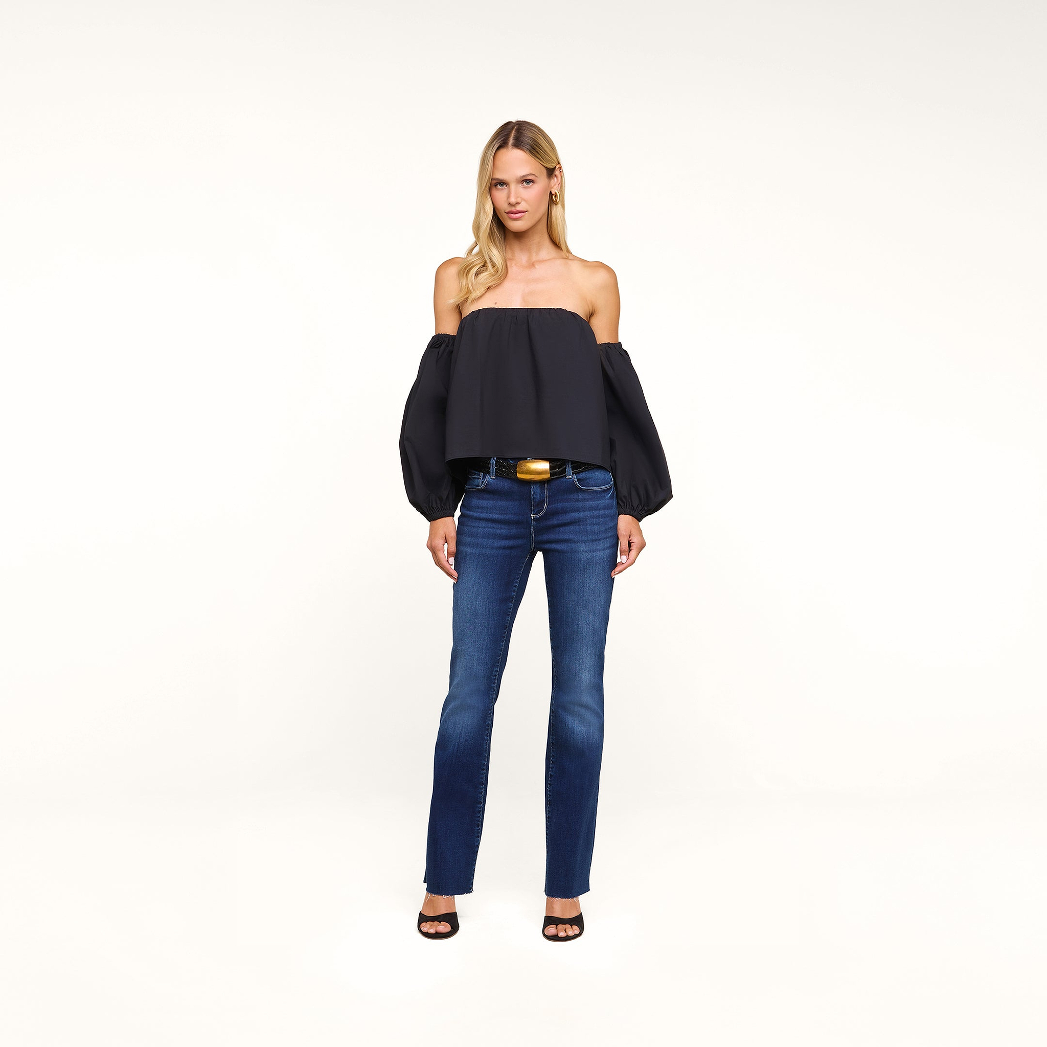 A woman stands against a white background wearing the Sarai Off-The-Shoulder Top—an organic cotton-blend black piece featuring puffed sleeves—paired with blue jeans, black open-toe heels, and a wide belt with a gold buckle.