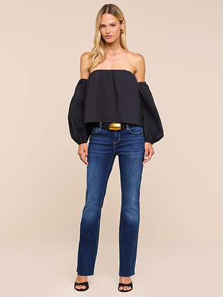 A woman stands against a plain background, wearing the Sarai Off-The-Shoulder Top with long puff sleeves crafted from organic cotton, paired with blue jeans, a black belt featuring a gold buckle, and black heels. Her long blonde hair frames her face as she maintains a neutral expression.