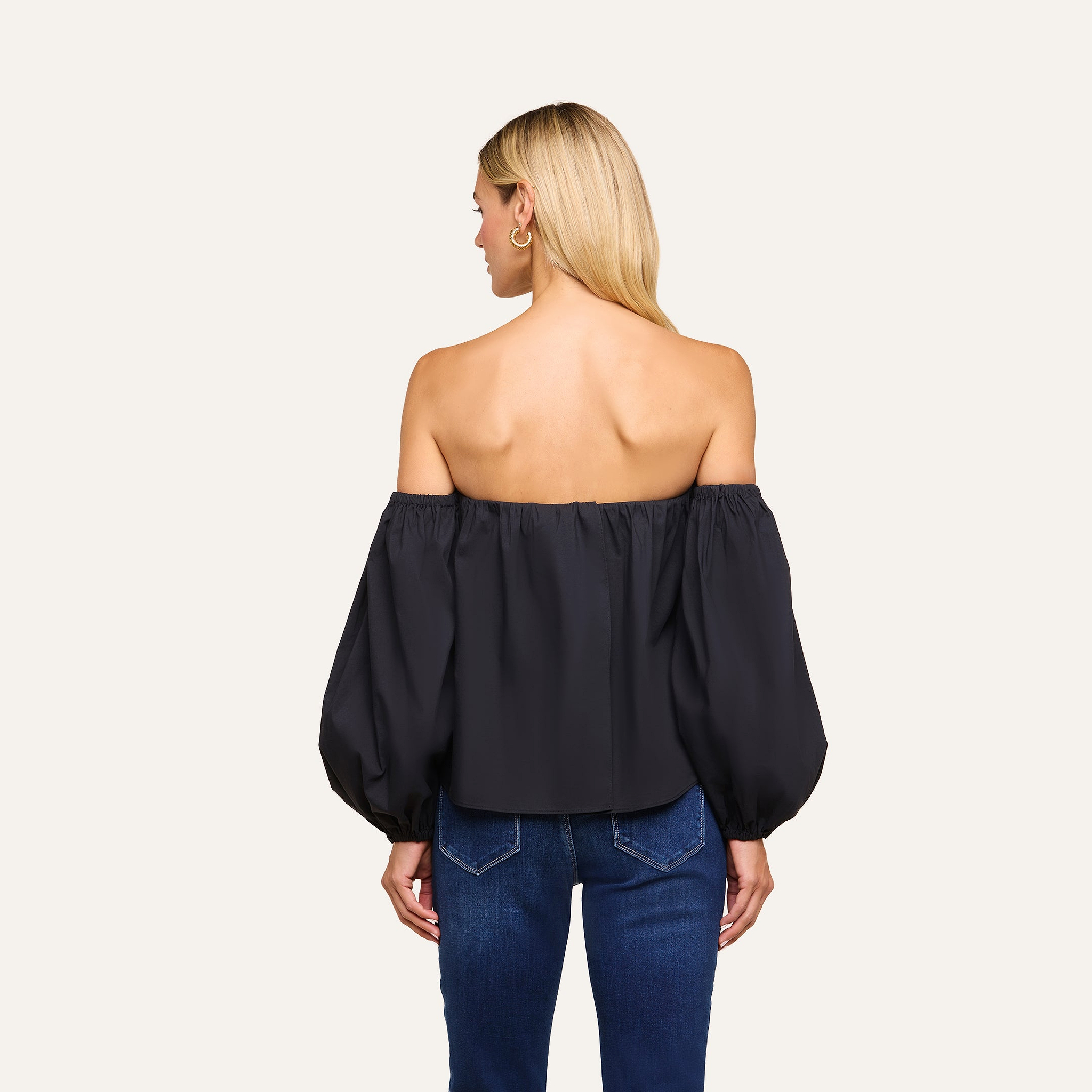 A woman with long blonde hair is seen from the back wearing the Sarai Off-The-Shoulder Top, featuring puffy sleeves and crafted from an organic cotton-blend, paired with blue jeans against a plain light background.