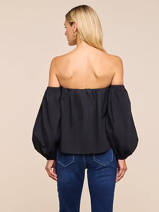 A woman with long blonde hair, seen from behind, wears the Sarai Off-The-Shoulder Top with puffy sleeves and blue jeans against a plain beige background. This stylish top features a built-in bra, combining comfort and elegance for an effortlessly chic look.