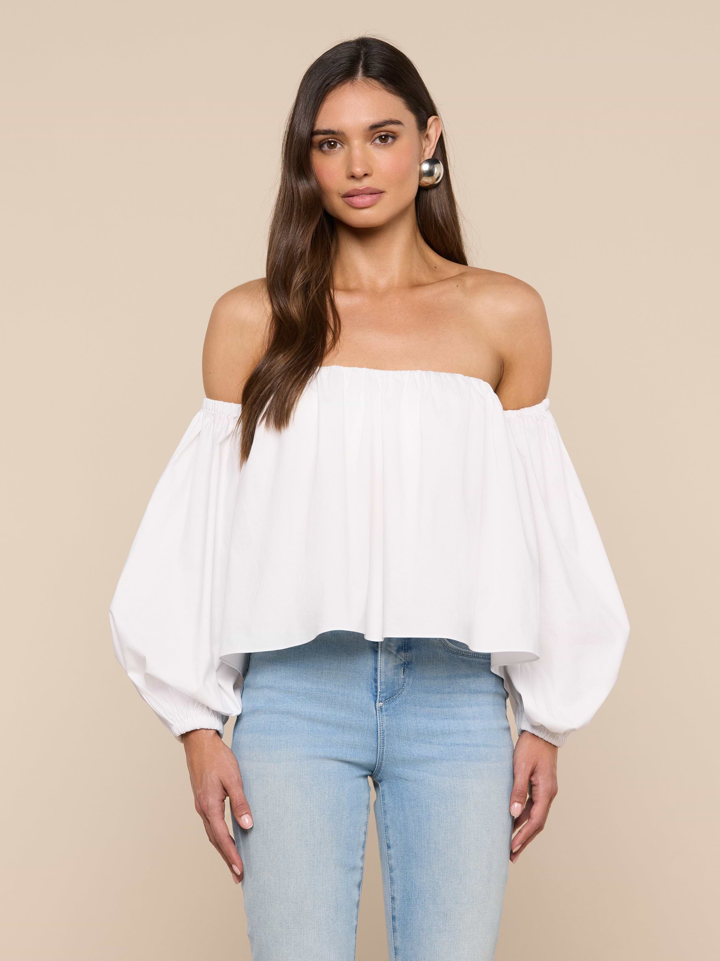 A woman stands against a plain light background wearing the Sarai Off-the-Shoulder Top—an off-the-shoulder, long-sleeve blouse made from organic cotton-blend fabric—and light blue jeans.