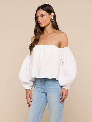 A woman with long brown hair and large hoop earrings stands against a beige background wearing the Sarai Off-the-Shoulder Top, a white organic cotton long-sleeve blouse, and light blue high-waisted jeans.