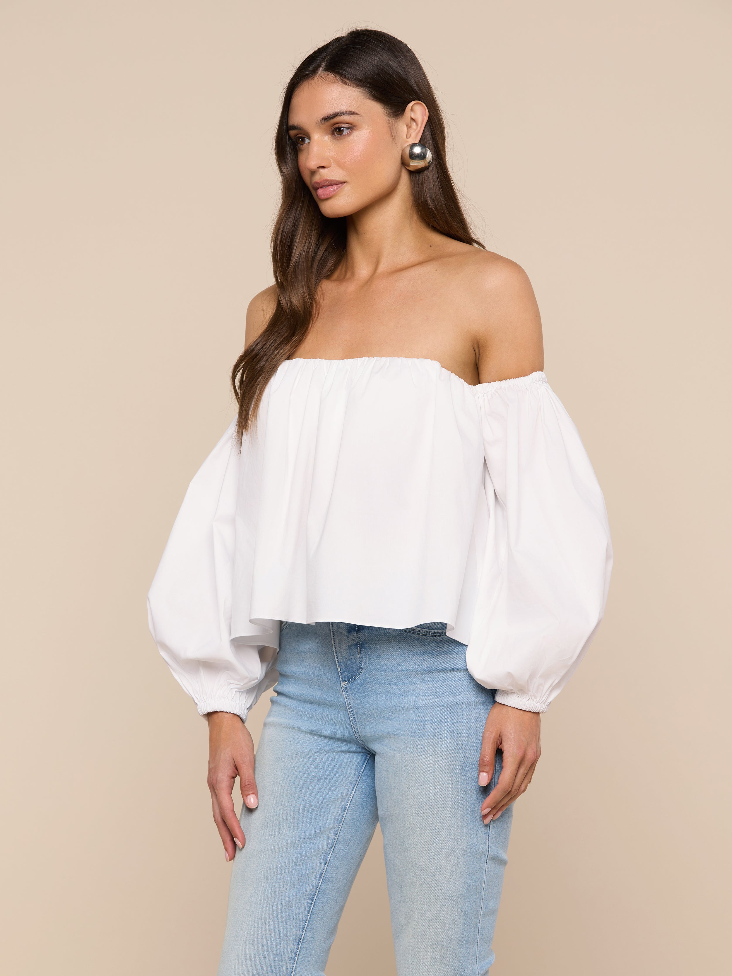 Wearing the Sarai Off-the-Shoulder Top—crafted from an organic cotton-blend with long puffed sleeves—a woman pairs it with light blue jeans and stands against a plain light background.