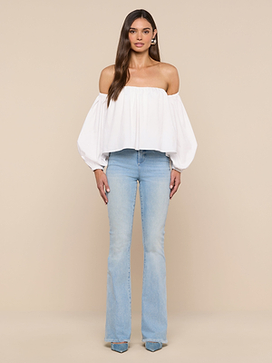 A woman poses against a beige background in the Sarai Off-the-Shoulder Top, a white long-sleeve blouse made from organic cotton, paired with light blue flared jeans and pointed-toe shoes.