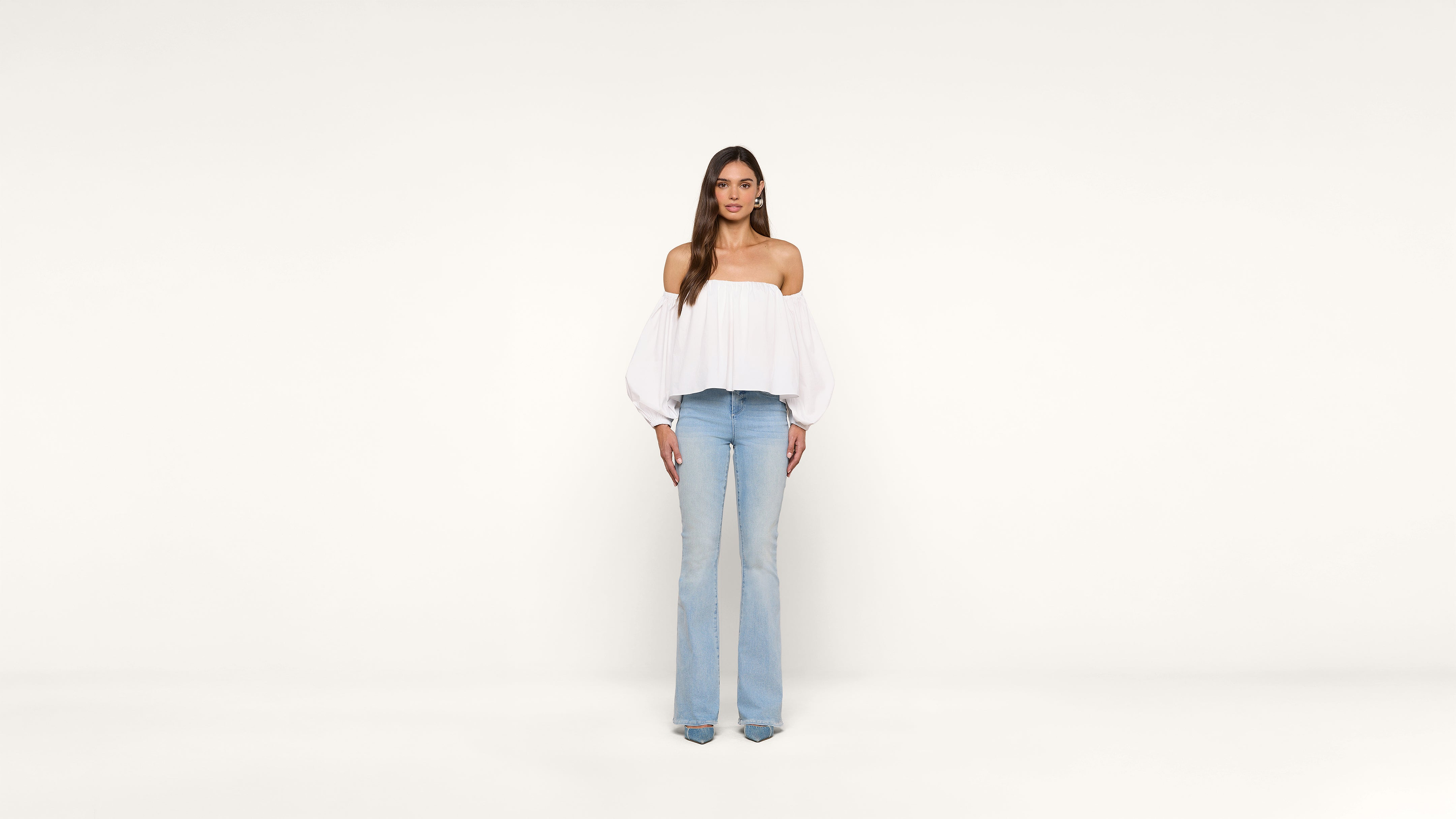 A woman stands facing forward against a plain white background, wearing a white off-the-shoulder top and light blue jeans.