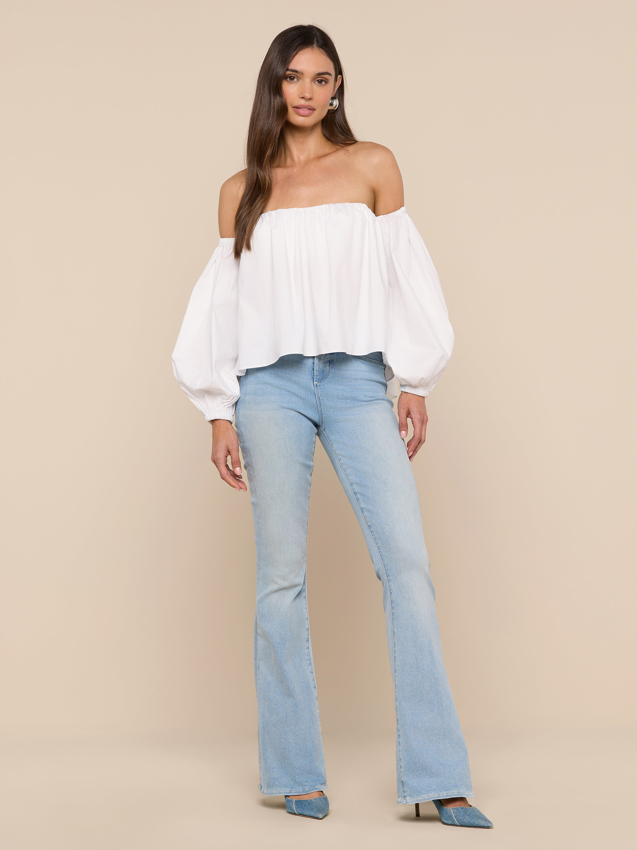 A woman stands against a white background wearing the Sarai Off-the-Shoulder Top, featuring billowy sleeves and crafted from an organic cotton-blend, paired with light blue flared jeans and blue pointed-toe shoes.