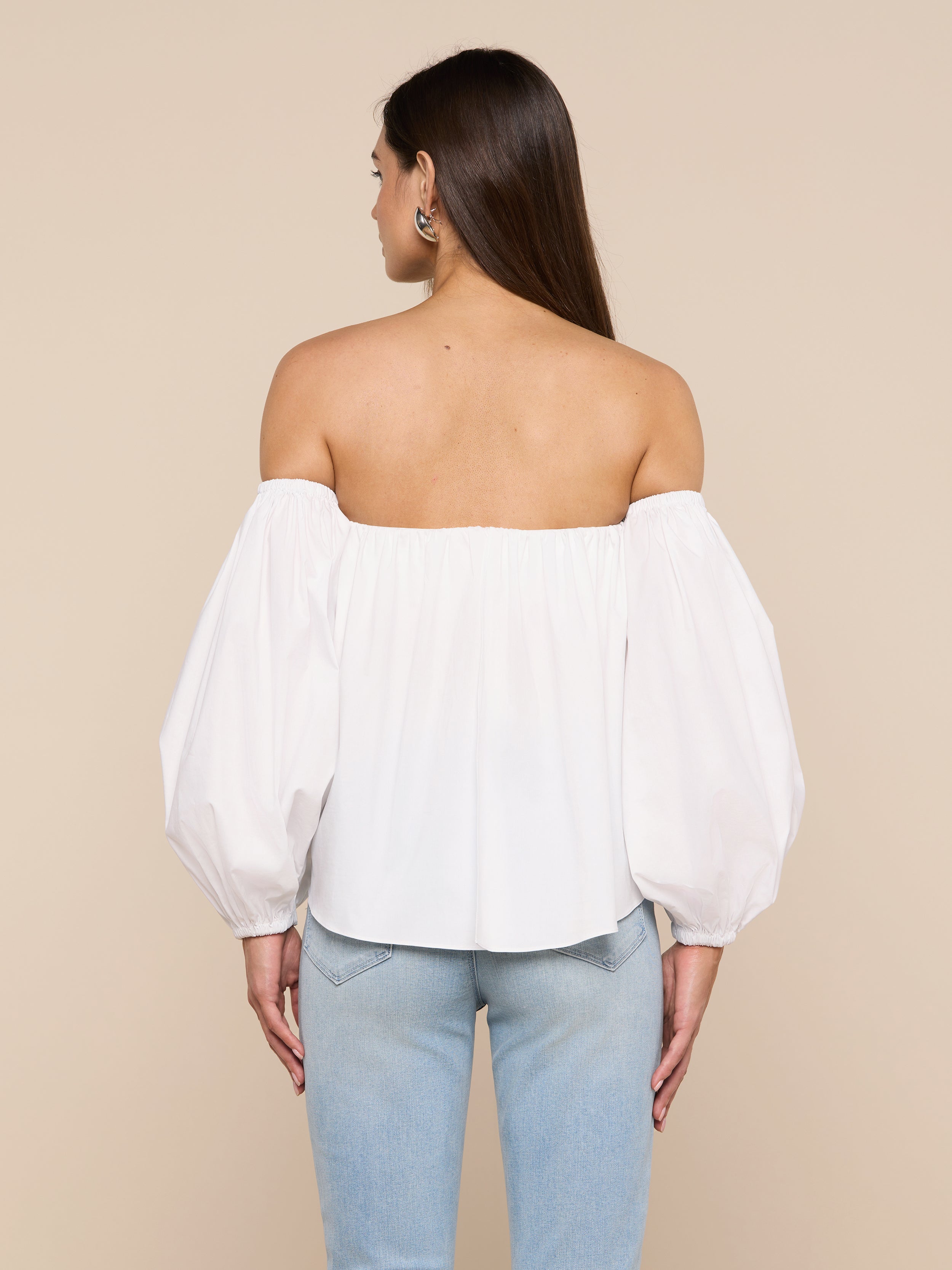A woman is seen from behind wearing the Sarai Off-the-Shoulder Top—a white blouse with puffy sleeves made from an organic cotton-blend—paired with light blue jeans. She has straight brown hair and stands against a plain light background.