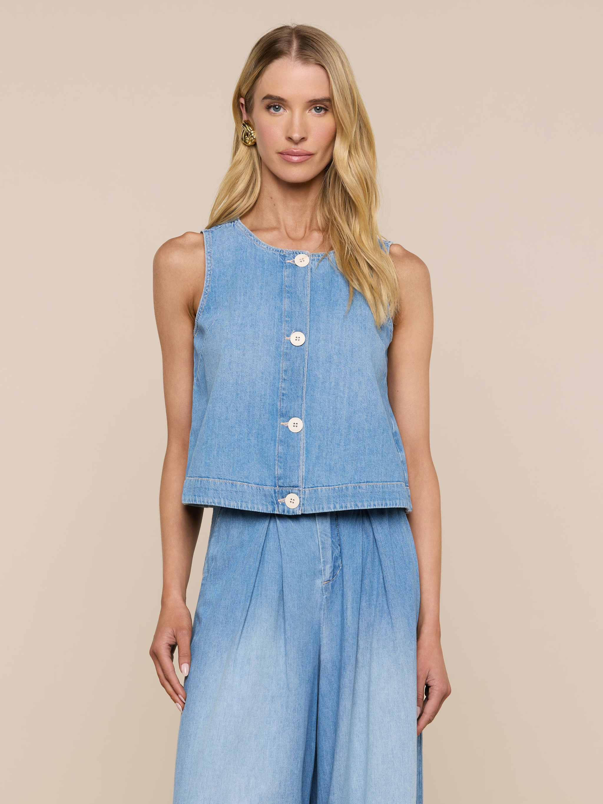 Una donna dai lunghi capelli biondi si staglia su uno sfondo beige, indossando il Foster Top - un capo in denim senza maniche e dal peso piuma - abbinato a pantaloni in denim a vita alta e a gamba larga con una silhouette ad A, guardando direttamente la telecamera.