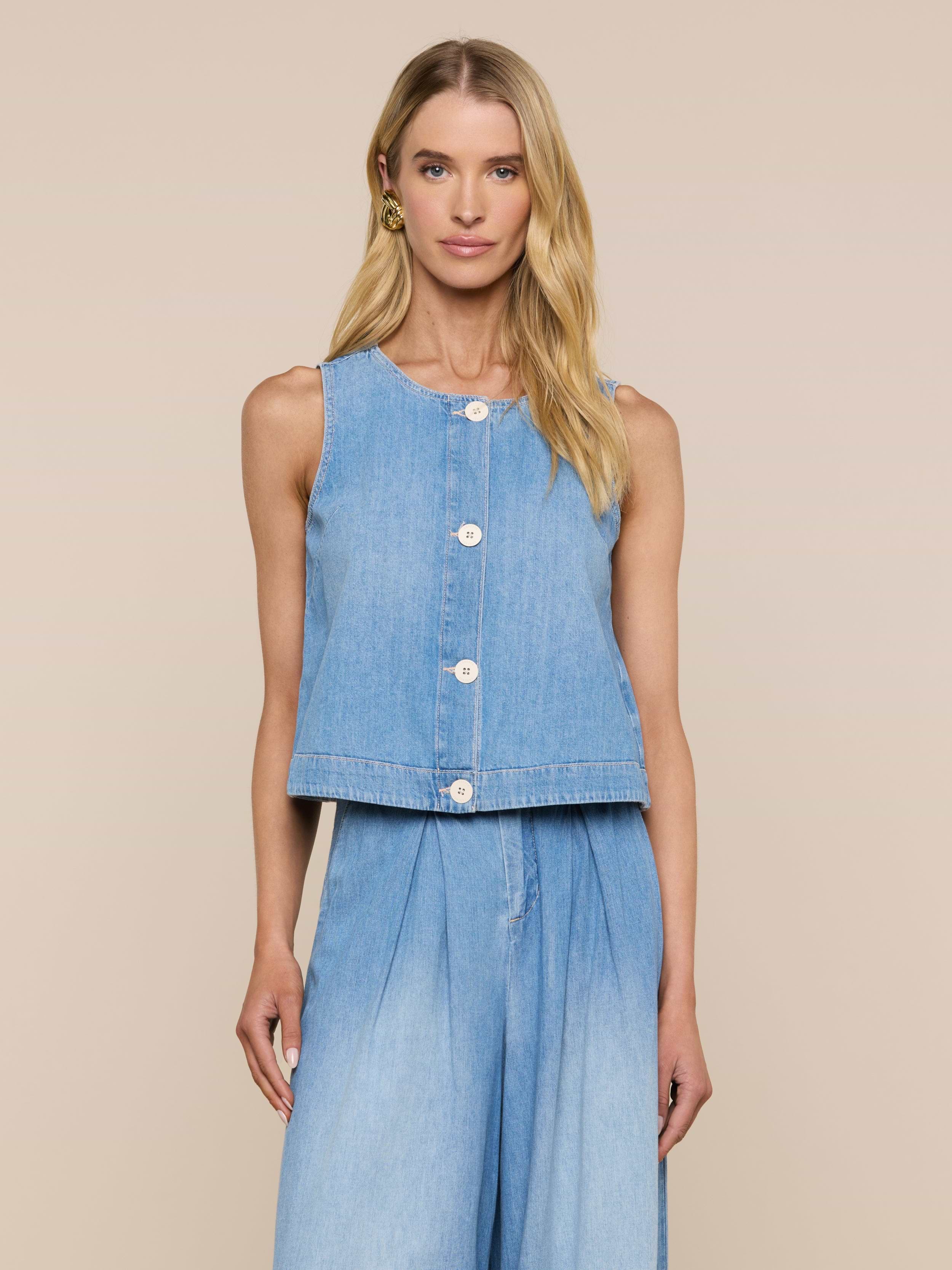 Una donna dai lunghi capelli biondi si staglia su uno sfondo beige, indossando il Foster Top - un capo in denim senza maniche e dal peso piuma - abbinato a pantaloni in denim a vita alta e a gamba larga con una silhouette ad A, guardando direttamente la telecamera.