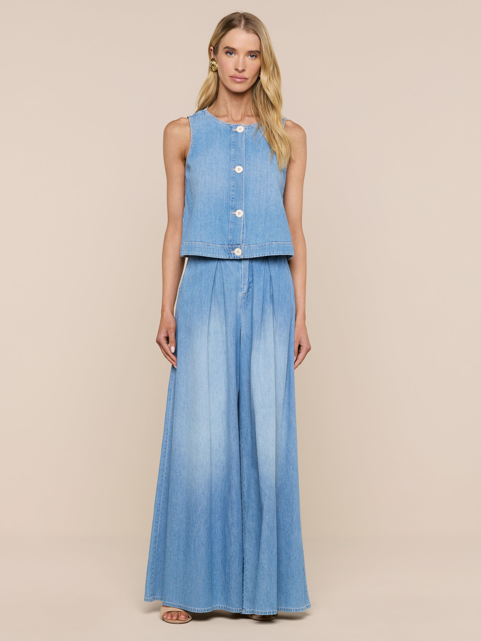 Una donna dai lunghi capelli biondi si staglia su uno sfondo beige, indossando il Foster Top senza maniche in denim leggero come una piuma e i pantaloni in denim a vita alta a gamba larga abbinati a sandali.