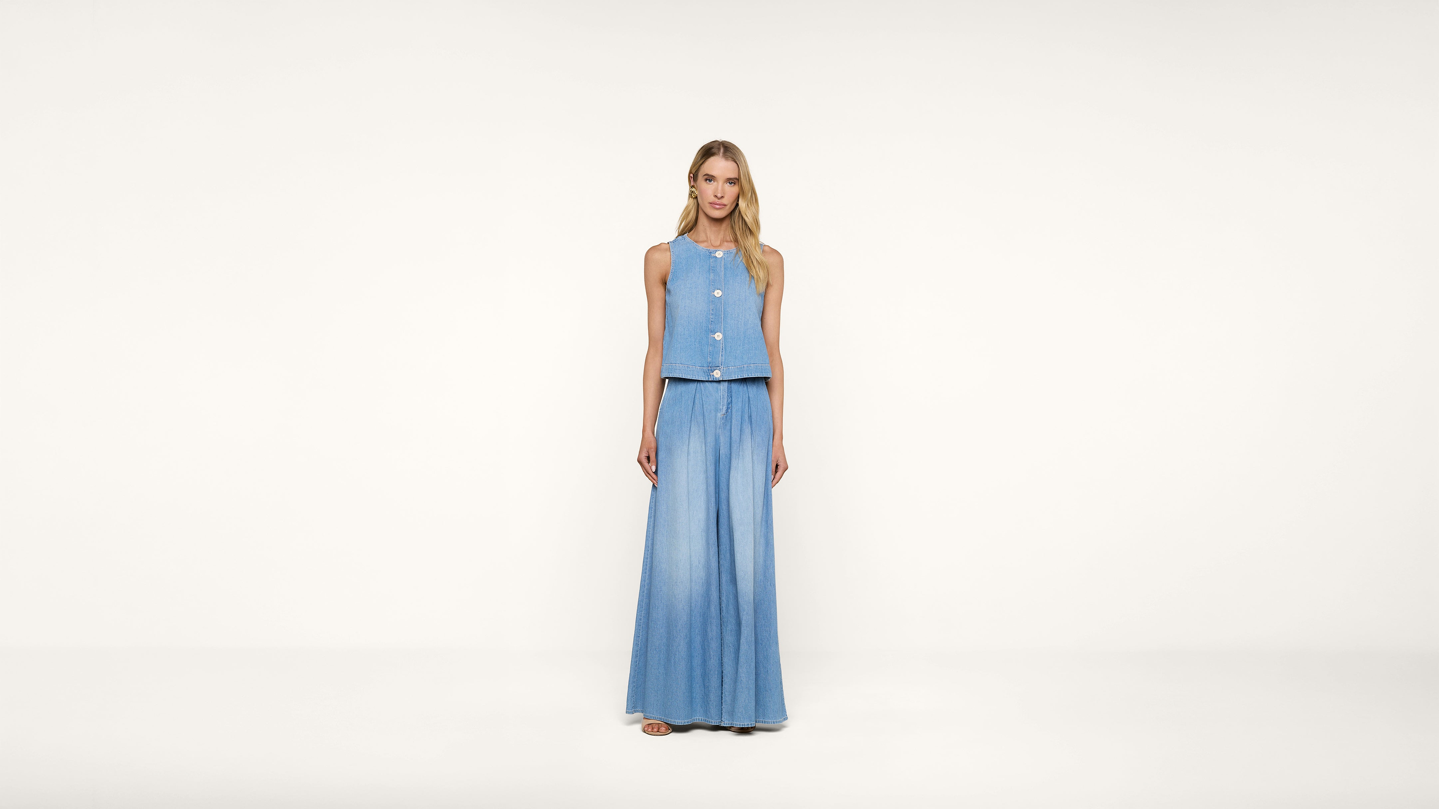 A woman stands against a plain white background, wearing a sleeveless blue button-up top and a long, matching blue skirt.