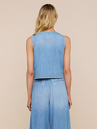 Una donna dai lunghi capelli biondi è ritratta di spalle, con indosso il Foster Top, un gilet in denim senza maniche e peso piuma, abbinato a pantaloni in denim a gamba larga, su uno sfondo beige.
