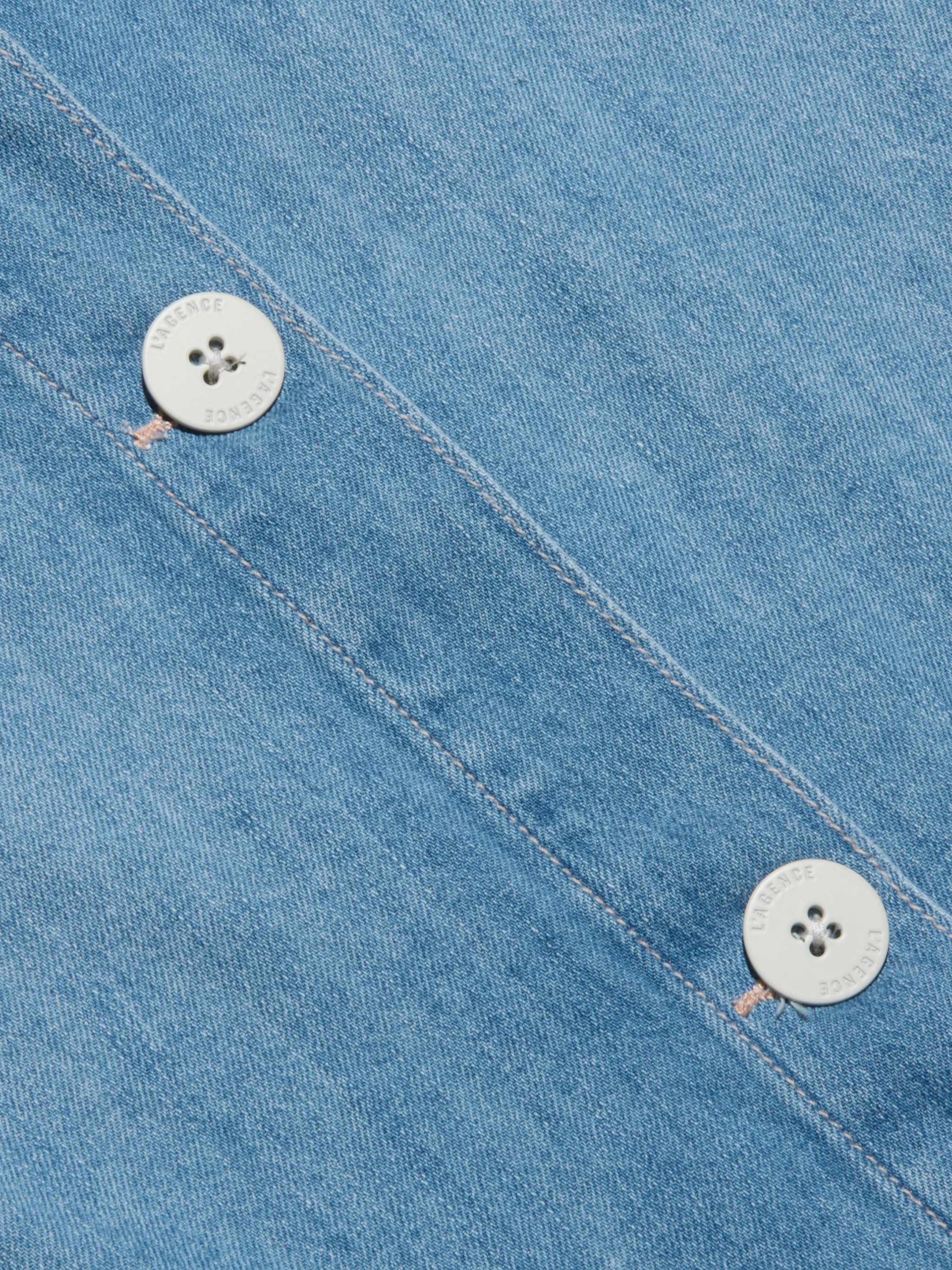 Primo piano di un tessuto denim blu con due bottoni bianchi cuciti lungo una cucitura diagonale. I bottoni hanno quattro fori e un sottile testo inciso intorno ai bordi. La texture del denim è chiaramente visibile.