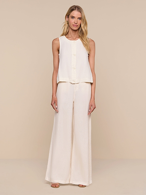A woman with long blonde hair and a neutral expression poses against a beige background, wearing the Foster Linen Top and matching wide-leg pants with open-toe sandals for an effortless summer look.