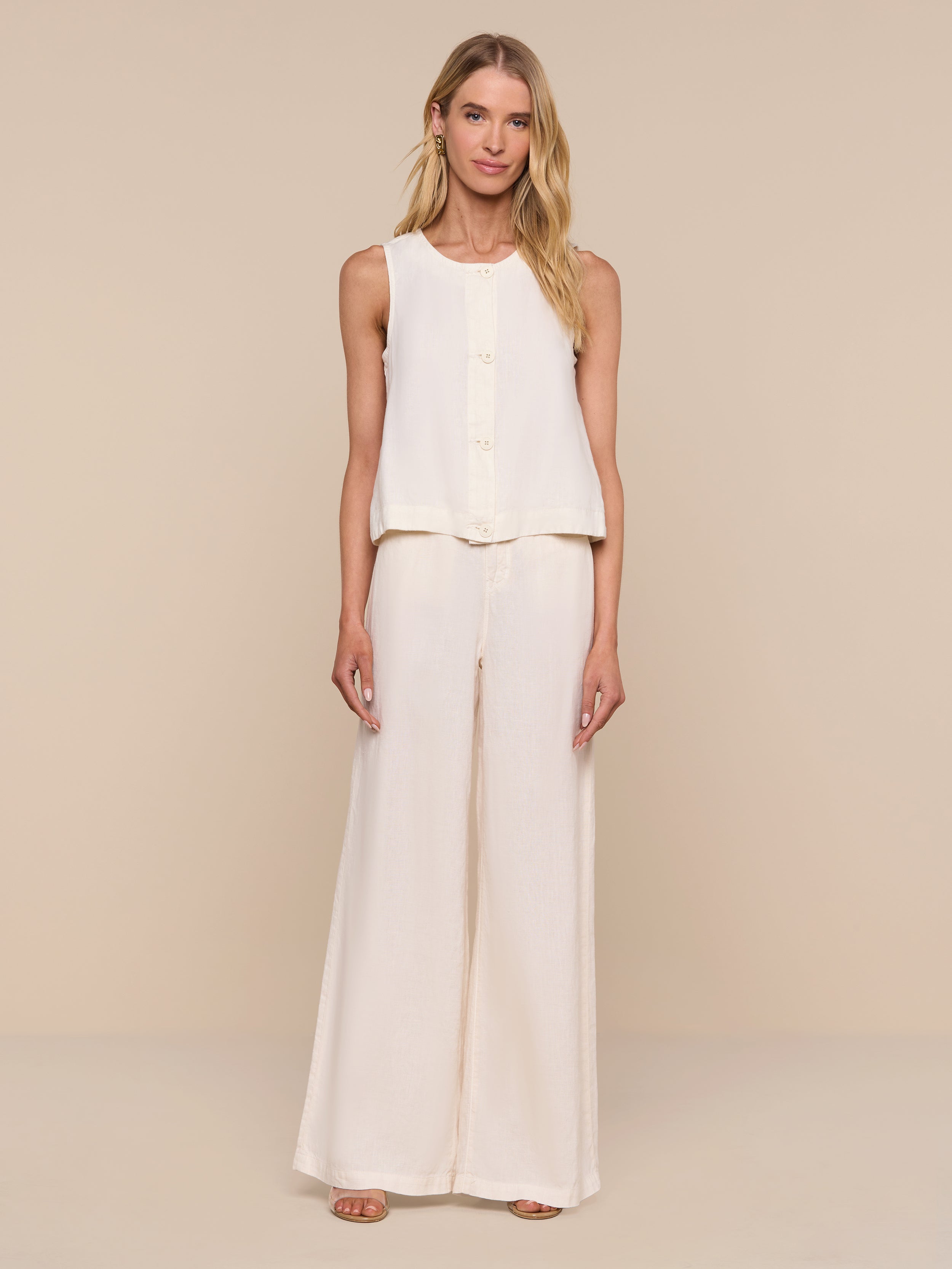 A woman stands against a plain white background, wearing the sleeveless Foster Linen Top with an A-line silhouette and matching wide-leg pants, paired with open-toe shoes.