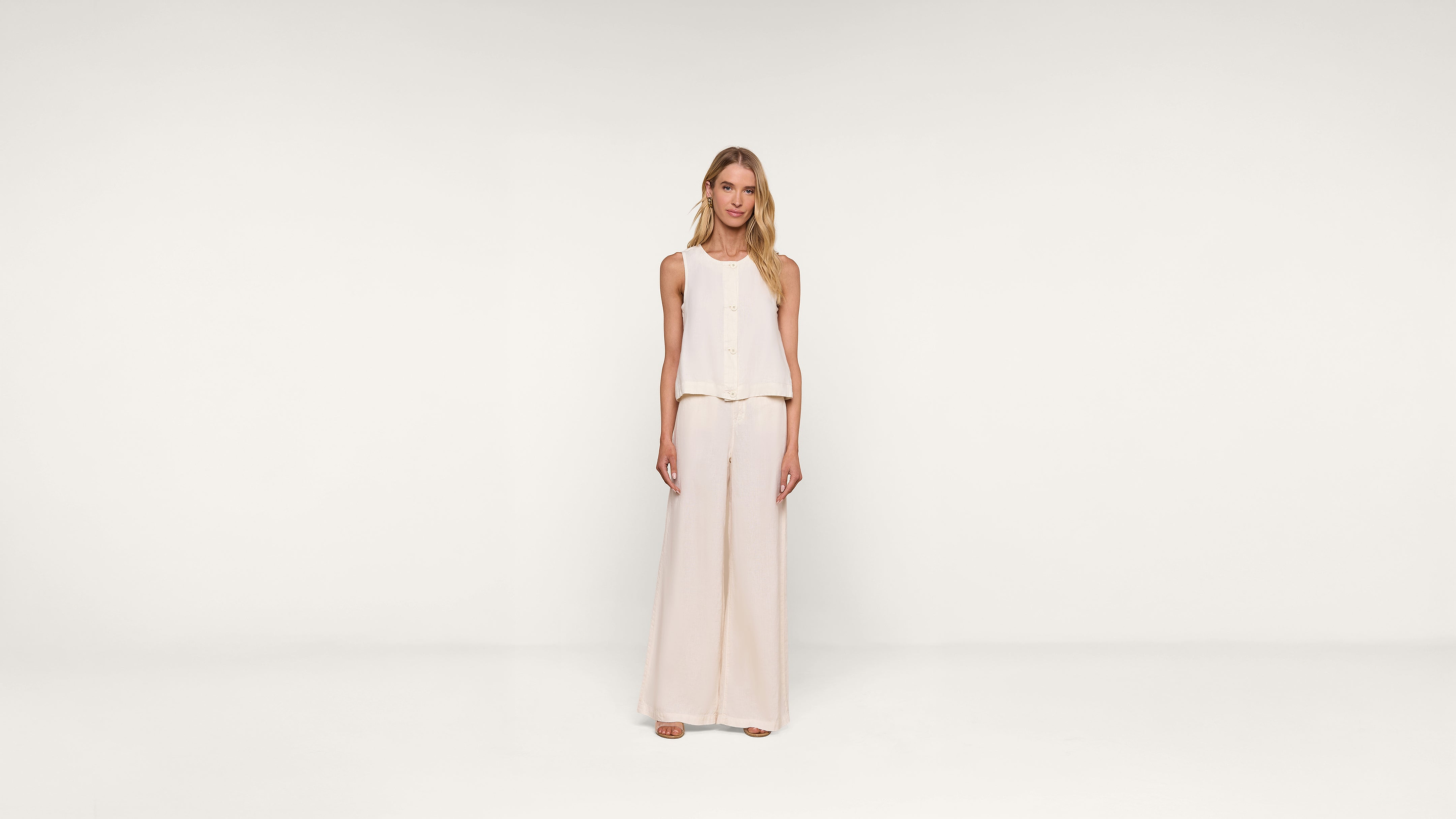A woman stands facing forward, wearing a sleeveless, light beige top and matching wide-leg pants against a plain white background.