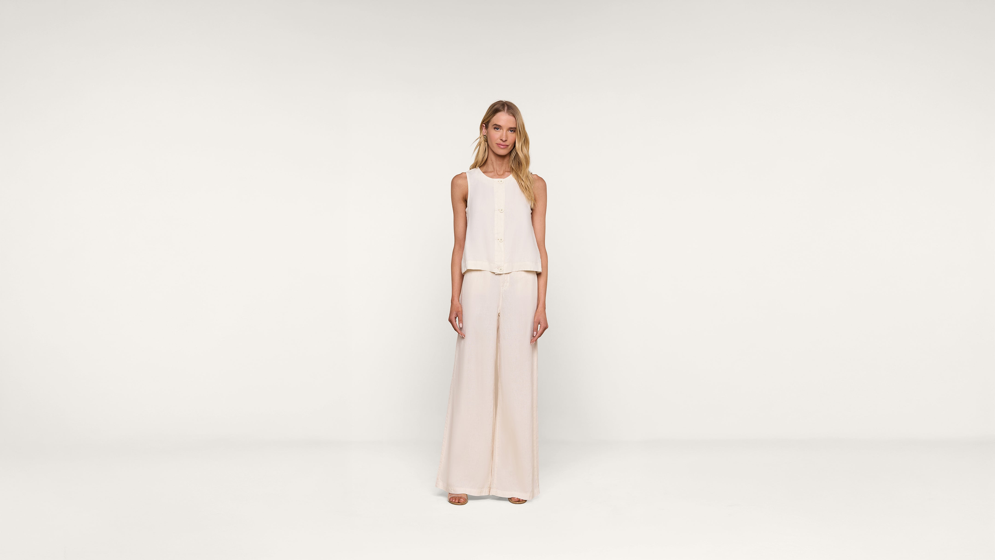 A woman stands facing forward, wearing a sleeveless, light beige top and matching wide-leg pants against a plain white background.