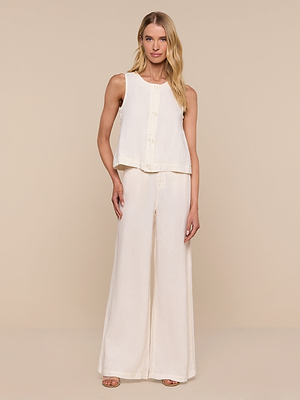 A woman with long blonde hair stands against a beige background wearing the Foster Linen Top and matching wide-leg pants with sandals, exuding a relaxed and confident vibe.