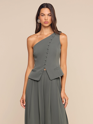 A woman wears the ash olive Zinnia One-Shoulder Top, showcasing sculptural tailoring and a diagonal row of buttons across the front, paired with a flared skirt, as she stands against a plain beige background.