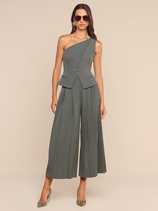 Wearing sunglasses, the woman stands against a neutral background in the ash olive Zinnia One-Shoulder Top styled with sculptural tailoring, decorative buttons, wide-leg cropped pants, and pointed heels.